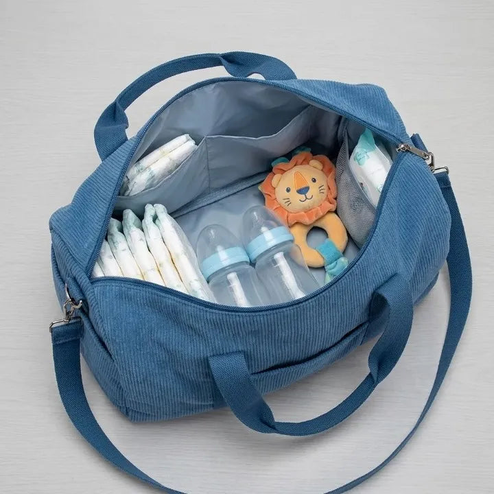 Sac à langer en velours côtelé bleu pétrole ouvert vue du dessus révélant son intérieur spacieux : couches, biberons transparents, hochet lion et lingettes bébé organisés dans les compartiments
