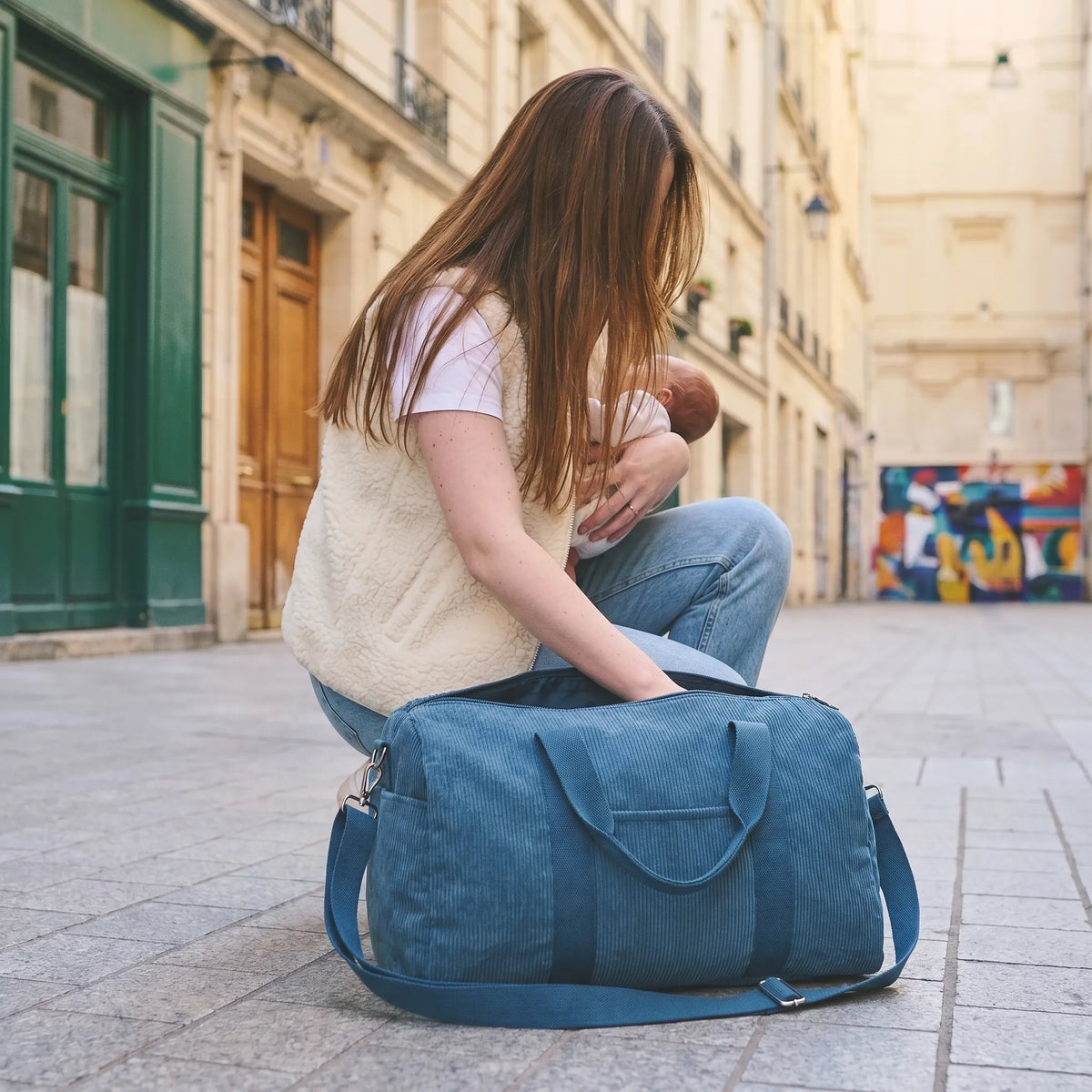 Maman aux longs cheveux châtain accroupie sur un trottoir parisien tenant son bébé contre elle tout en cherchant dans son sac à langer en velours côtelé bleu pétrole posé au sol