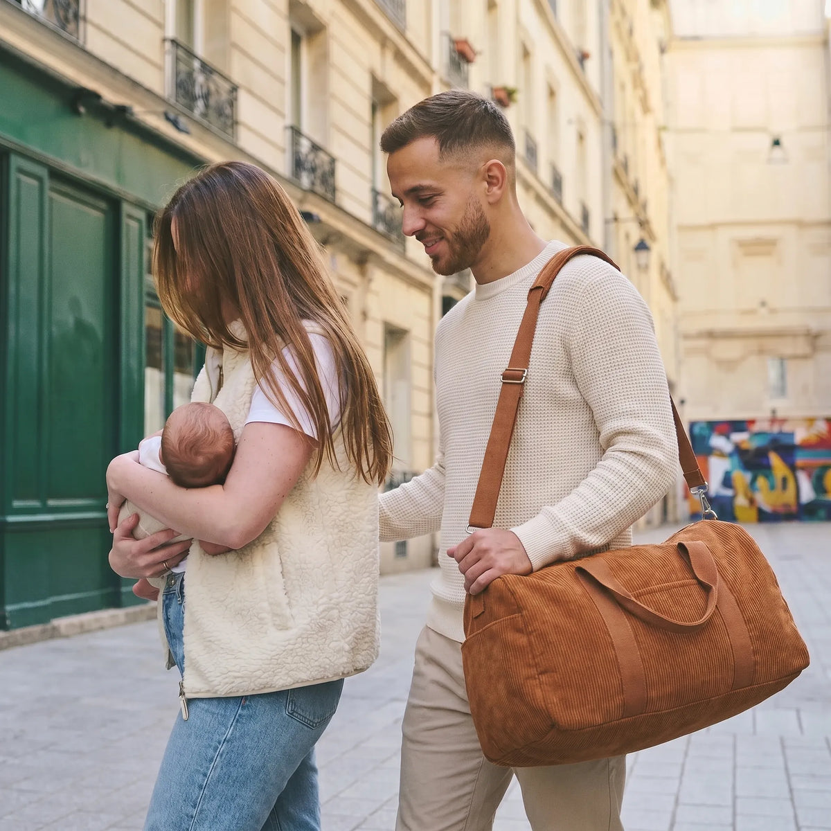 Couple de jeunes parents marchant dans une rue parisienne avec leur nouveau-né, le papa portant un sac à langer en velours côtelé caramel en bandoulière