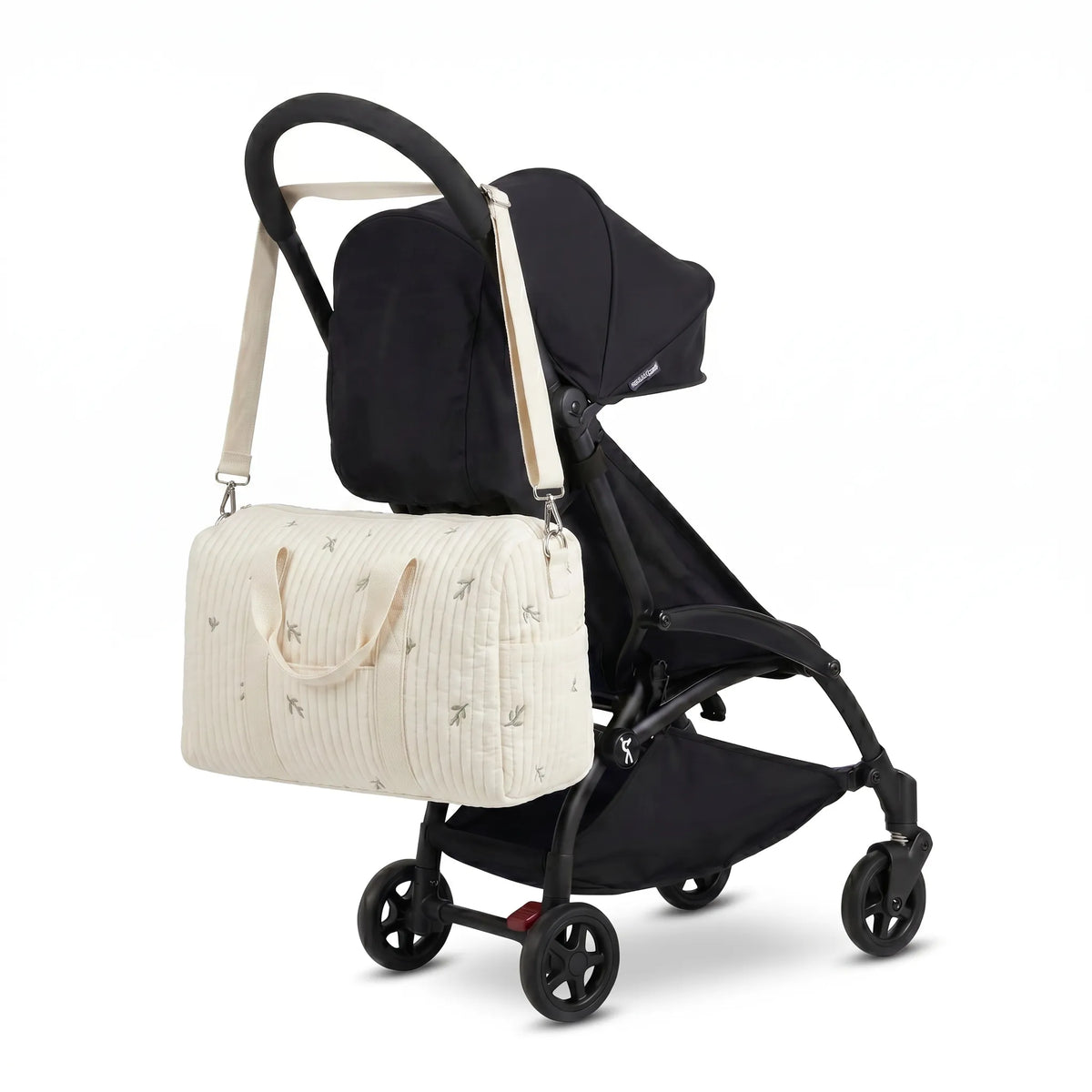 Sac maternité format polochon en gaze de coton matelassée écru à petit motif feuillage avec bandoulière accroché au guidon d'une poussette compacte noire de type Yoyo