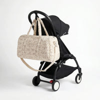 Sac maternité format polochon matelassé beige à imprimé fleuri avec bandoulière accroché au guidon d'une poussette compacte noire de type Yoyo