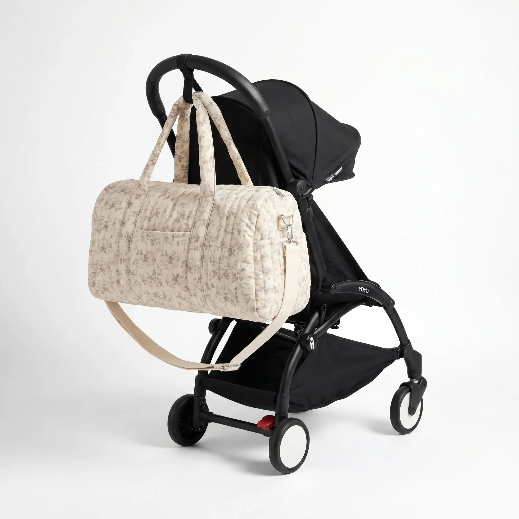 Sac maternité format polochon matelassé beige à imprimé fleuri avec bandoulière accroché au guidon d'une poussette compacte noire de type Yoyo