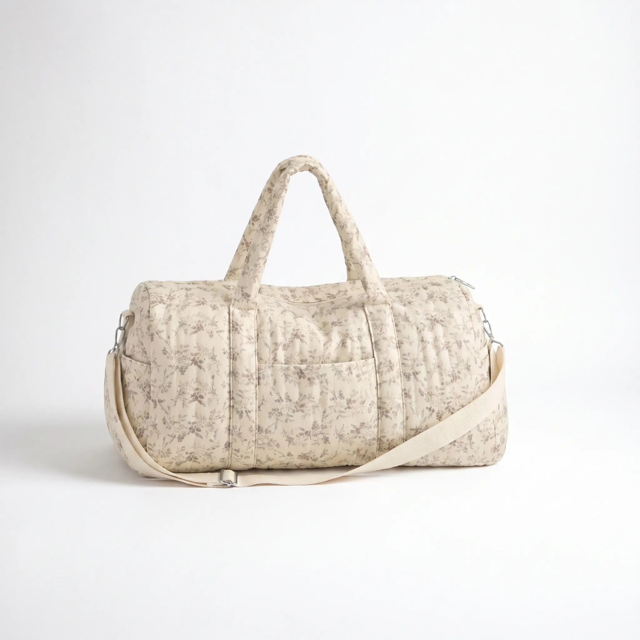 Sac maternité format polochon matelassé beige à imprimé fleuri délicat avec poignées, bandoulière ajustable et poche frontale sur fond clair