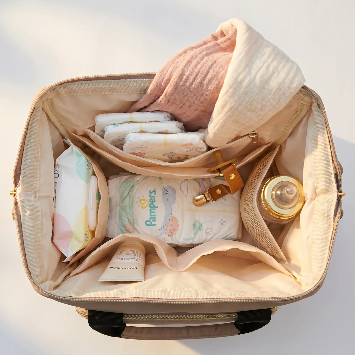 Vue plongeante sur l'intérieur d'un sac à dos à langer beige rempli avec couches Pampers, paquet de lingettes, crème de change, biberon dans la poche isotherme, attache-tétine et lange en gaze de coton rose