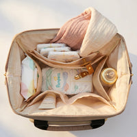 Vue plongeante sur l'intérieur d'un sac à dos à langer beige rempli avec couches Pampers, paquet de lingettes, crème de change, biberon dans la poche isotherme, attache-tétine et lange en gaze de coton rose