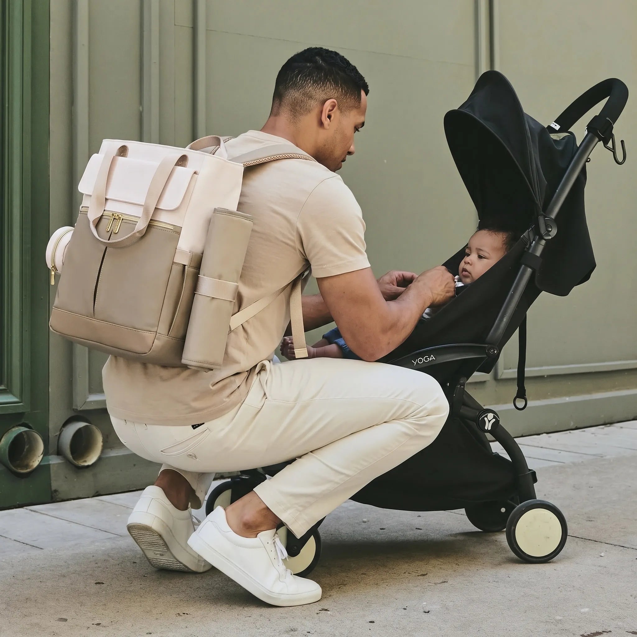 Un homme portant un sac à dos à langer moderne beige et crème, accroupi à côté d'un bébé dans une poussette noire YOYO pour ajuster ses sangles.